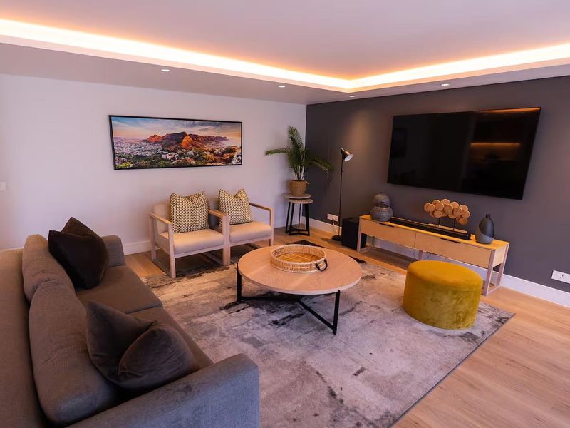 A modern, well-lit living room featuring hardwood flooring, a dark accent wall, and sophisticated recessed cove lighting.