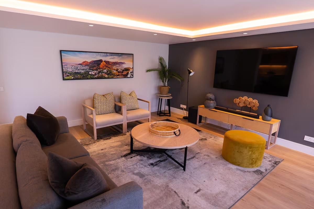 A modern, well-lit living room featuring hardwood flooring, a dark accent wall, and sophisticated recessed cove lighting.