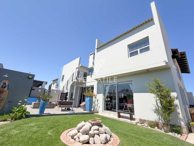 Rear view of the property featuring a paved patio area, artificial lawn, stone landscaping, and an external staircase leading to an upper balcony.