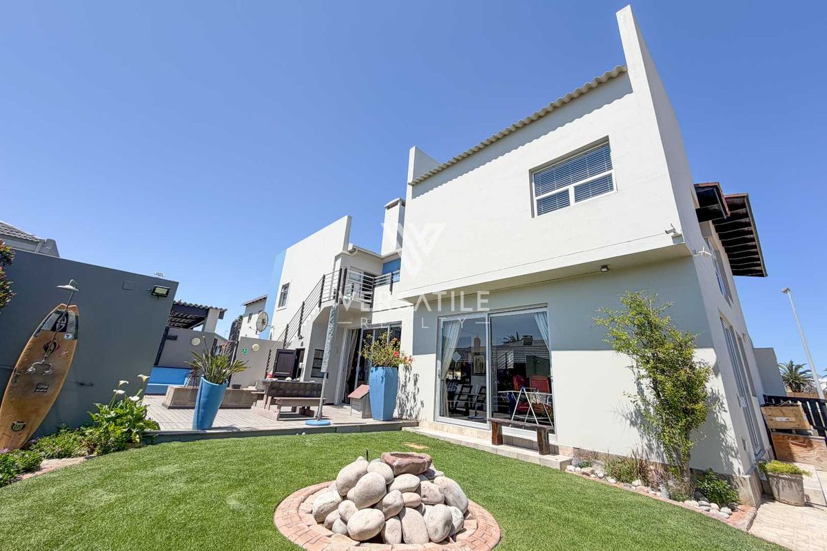 Rear view of the property featuring a paved patio area, artificial lawn, stone landscaping, and an external staircase leading to an upper balcony.