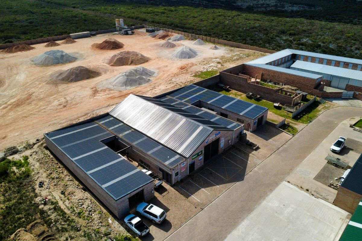 Aerial view of a commercial property featuring a large warehouse with extensive solar panel installation, paved parking, and a secure perimeter wall.