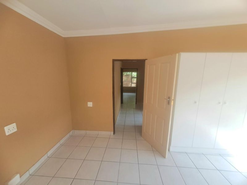 A clean, empty bedroom featuring neutral wall paint, white tiled flooring, and built-in cupboards, with a doorway leading to an adjacent hallway.