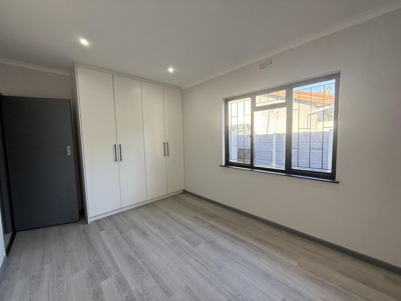 A bright, empty bedroom featuring modern laminate flooring, built-in white cupboards, and a window fitted with security bars.