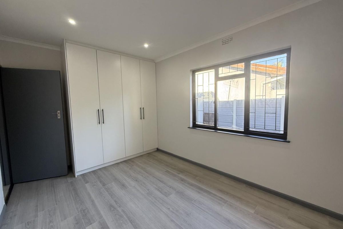 A bright, empty bedroom featuring modern laminate flooring, built-in white cupboards, and a window fitted with security bars.