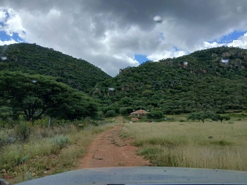 A scenic view of a rural farm property nestled at the base of a mountain range, accessed by a dirt track.
