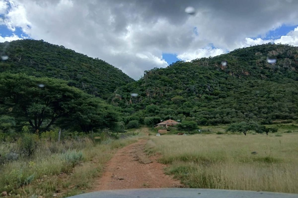 A scenic view of a rural farm property nestled at the base of a mountain range, accessed by a dirt track.