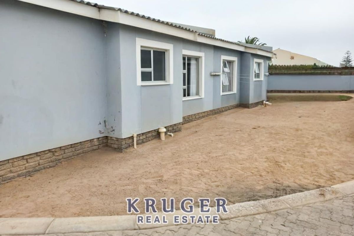 Rear exterior view of the townhouse featuring a plain facade, multiple windows, and an undeveloped sandy backyard area.
