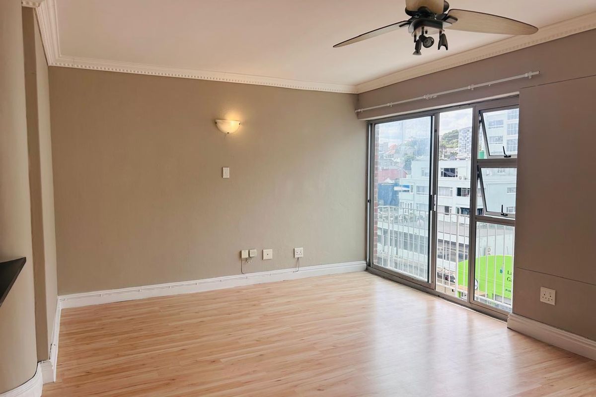 A bright, empty living room featuring laminate flooring, crown molding, a ceiling fan, and a large sliding glass door with urban views.