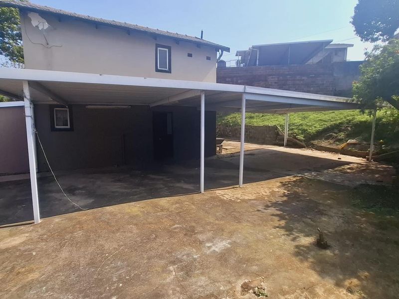 Rear exterior view featuring a large metal carport structure over a concrete driveway with visible wear to the building facade and paving.