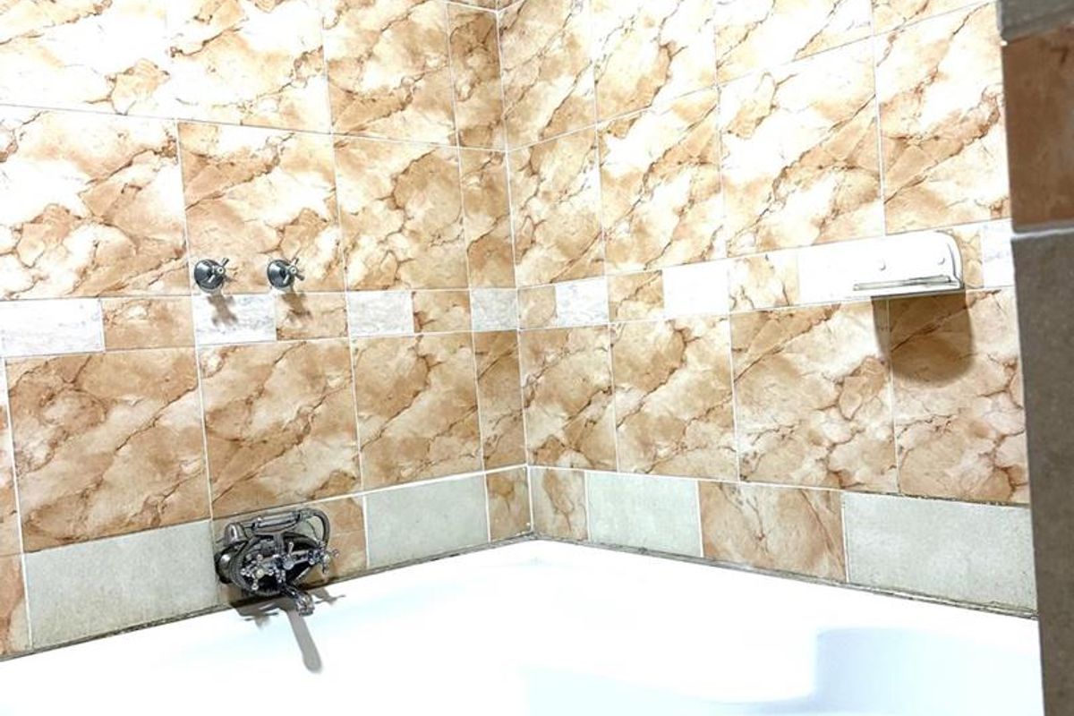 A corner bathtub featuring marble-effect wall tiling, a wall-mounted shower head, and chrome fixtures in a functional bathroom setting.