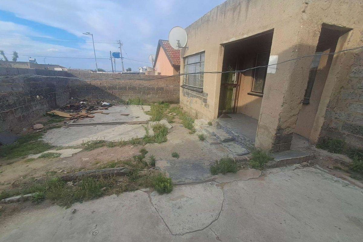 Rear exterior view of the property featuring a concrete patio area, perimeter boundary wall, and weathered building facade requiring maintenance.