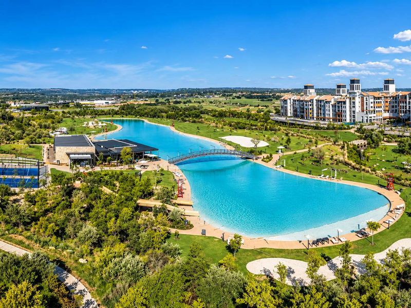 Aerial view of a residential estate featuring a large man-made lagoon, pedestrian bridge, sports courts, and extensive landscaped recreational grounds.
