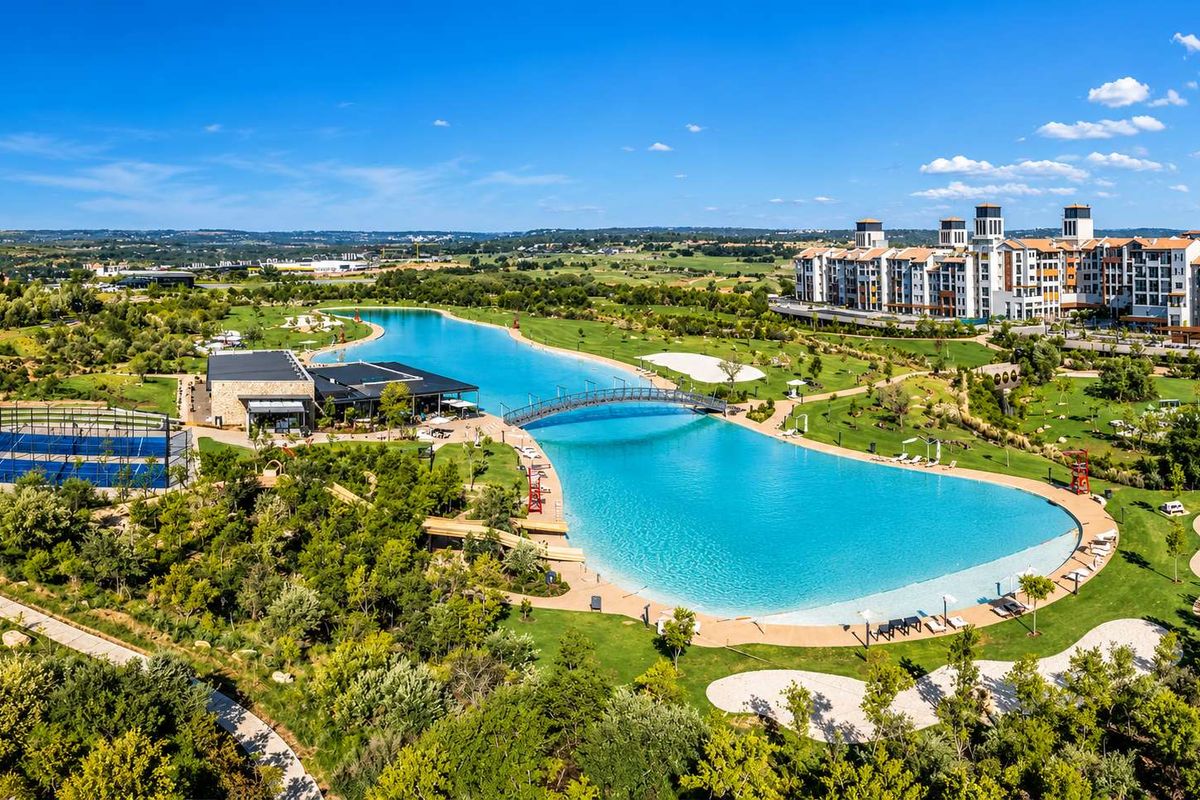Aerial view of a residential estate featuring a large man-made lagoon, pedestrian bridge, sports courts, and extensive landscaped recreational grounds.