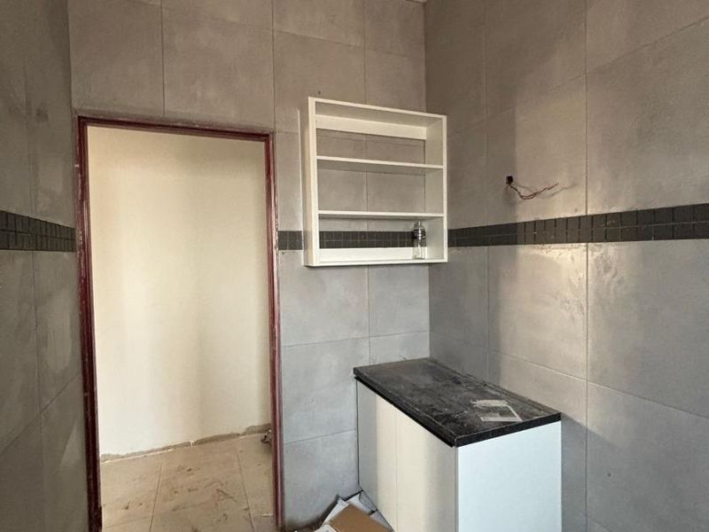 A compact scullery or utility area featuring wall-mounted and base cabinetry with stone countertops, finished with grey ceramic wall and floor tiles.