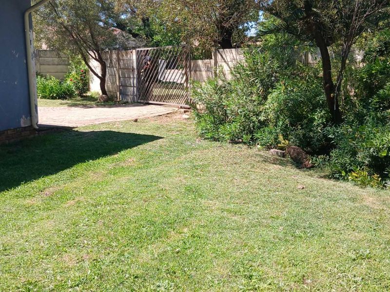 A view of the rear garden area featuring a lawn, paved driveway, metal gate, and boundary wall, with visible exterior wall maintenance required.