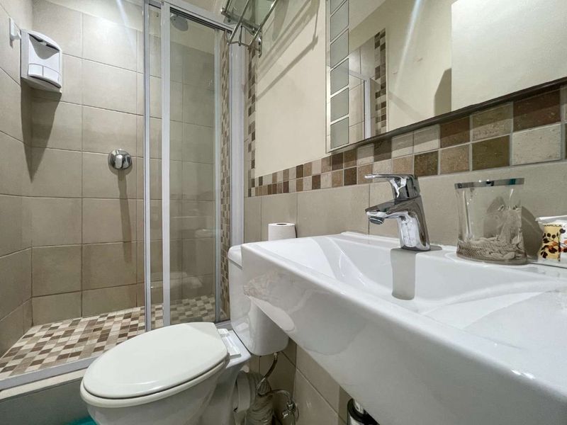 A compact, well-maintained bathroom featuring a glass-enclosed shower, modern vanity basin with chrome fixtures, and neutral tiling with decorative mosaic accents.