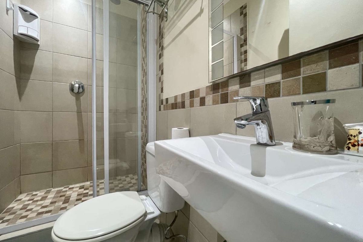 A compact, well-maintained bathroom featuring a glass-enclosed shower, modern vanity basin with chrome fixtures, and neutral tiling with decorative mosaic accents.