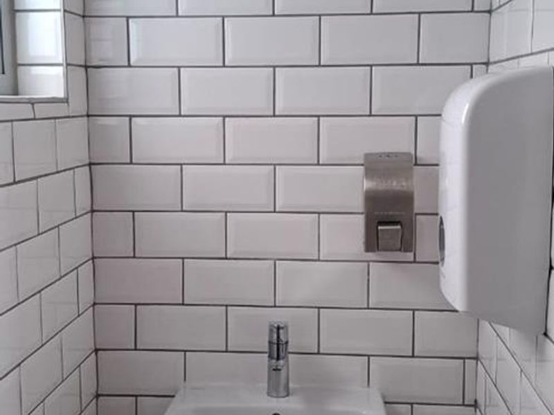 A clean, modern office restroom featuring white subway tile wall cladding, a wall-mounted basin with a chrome mixer tap, and wall-mounted dispensers.