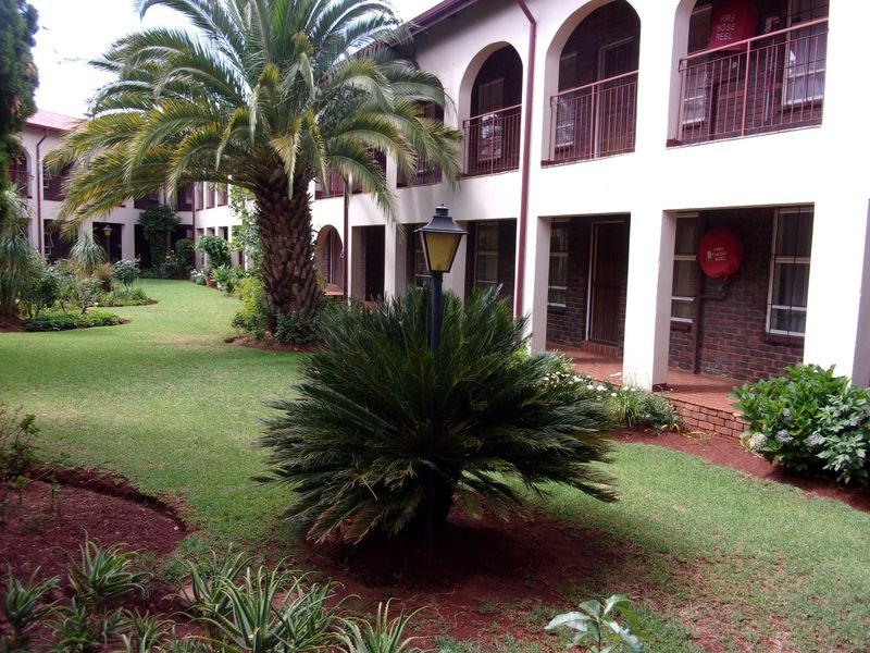 A well-maintained communal garden area featuring lush landscaping, mature palm trees, and arched walkways leading to apartment building entrances.