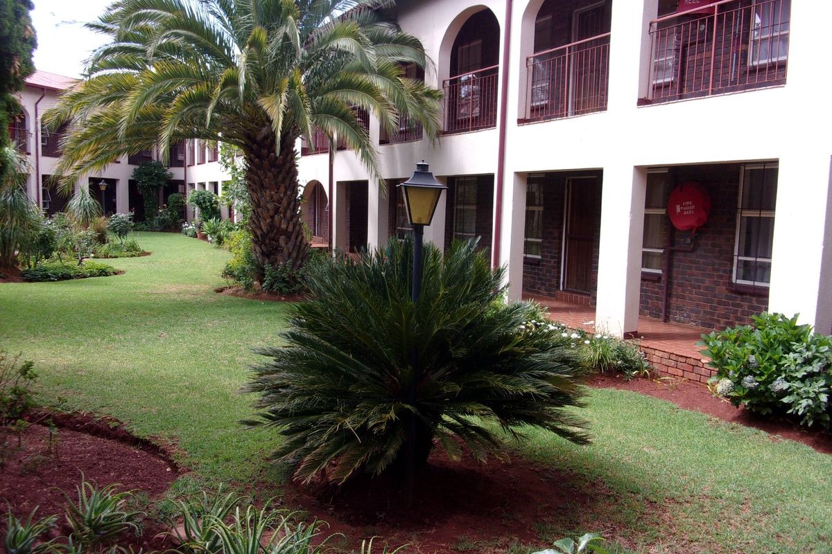 A well-maintained communal garden area featuring lush landscaping, mature palm trees, and arched walkways leading to apartment building entrances.