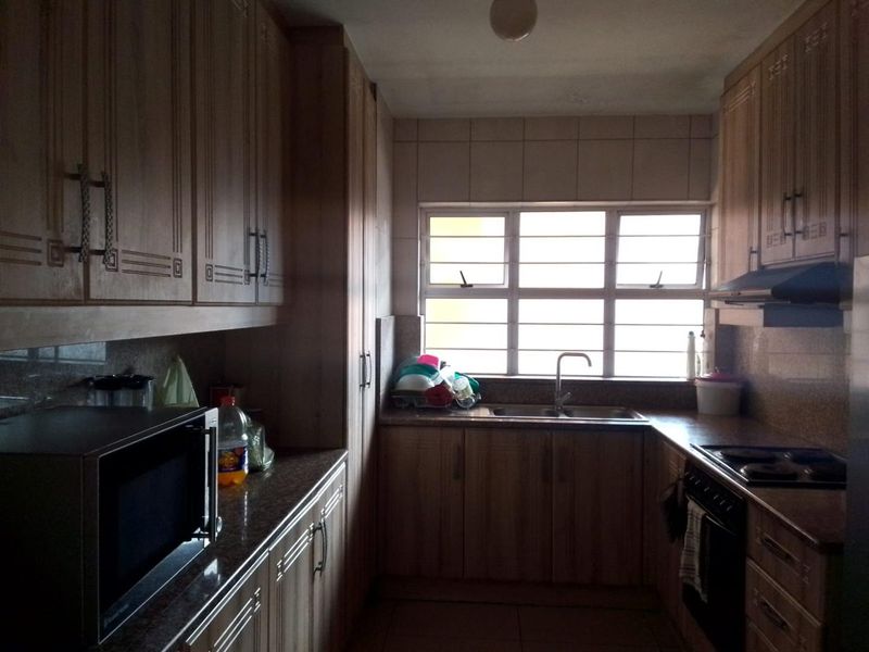 A functional galley-style kitchen featuring wood-finish cabinetry, laminate countertops, integrated oven and hob, and a window with security bars.