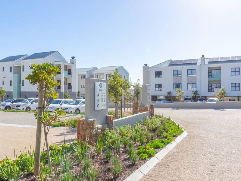 A well-maintained apartment complex exterior featuring paved parking areas, landscaped garden beds, and clear building signage under a bright sky.