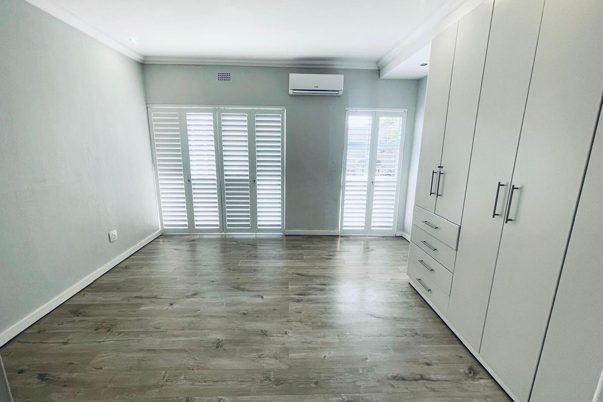 A bright, empty bedroom featuring modern laminate flooring, extensive built-in cupboards, air conditioning, and white plantation shutters.