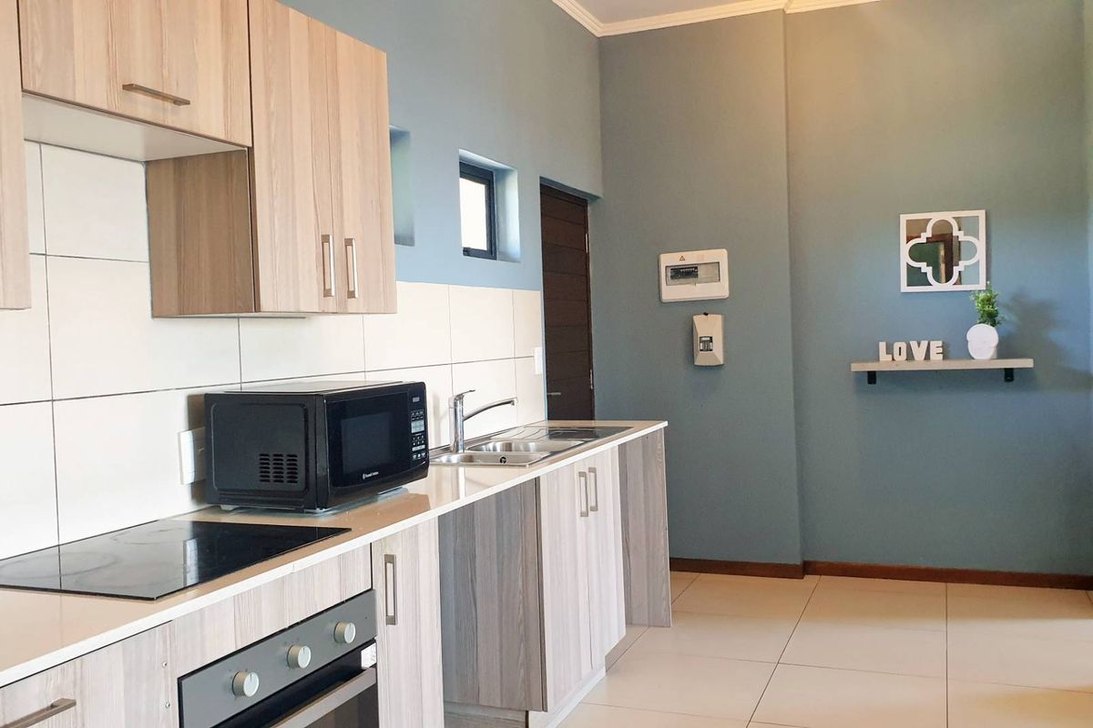 A functional kitchen featuring light wood-grain cabinetry, integrated electric hob and oven, stainless steel sink, and neutral tiled flooring.
