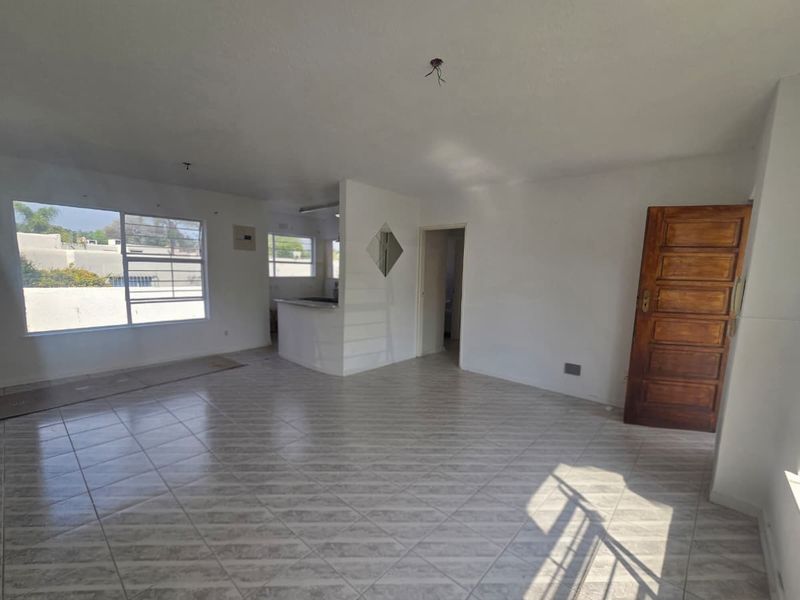 An empty open-plan living and kitchen area featuring tiled flooring, a breakfast bar, and natural light through large windows.