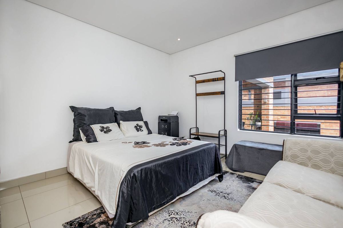A well-maintained bedroom featuring neutral tiled flooring, recessed ceiling lighting, and a large window with fitted security bars and a roller blind.