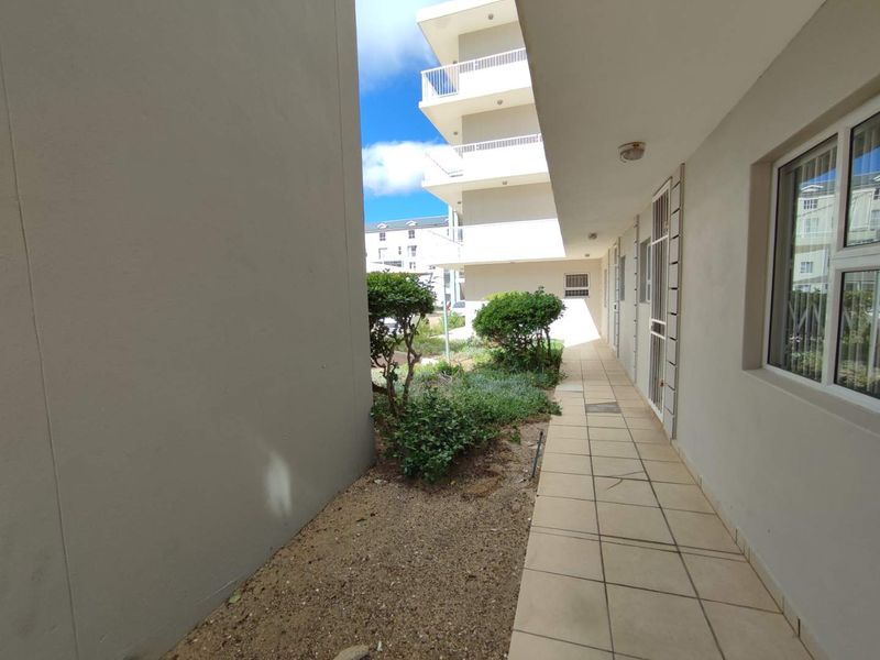 A paved exterior walkway alongside the apartment building, featuring security gates, aluminium window frames, and access to a small landscaped garden area.