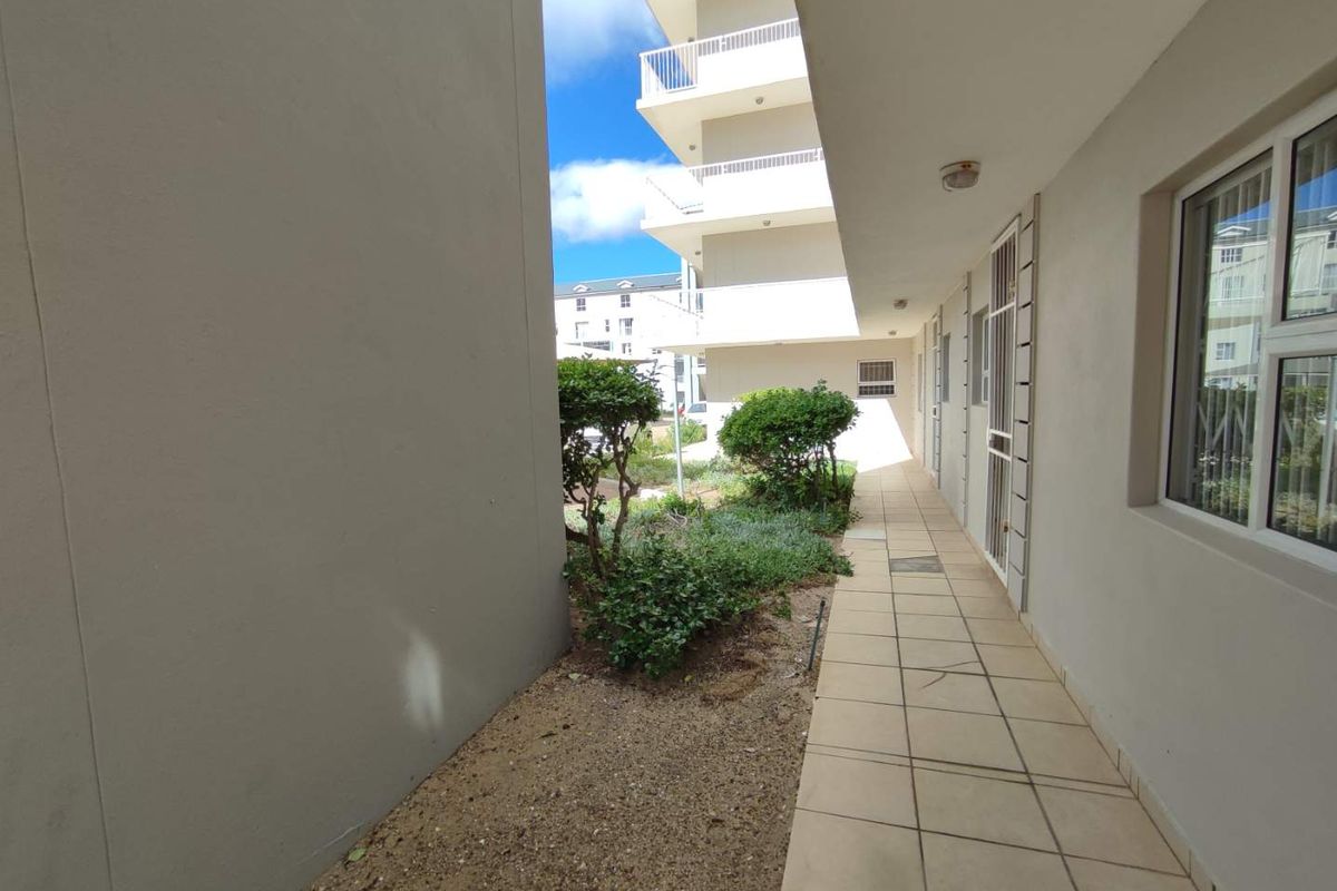 A paved exterior walkway alongside the apartment building, featuring security gates, aluminium window frames, and access to a small landscaped garden area.