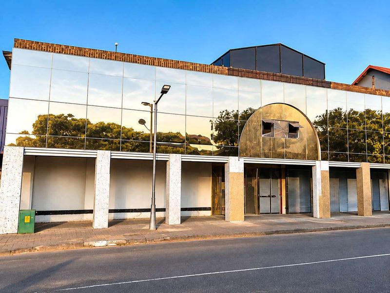 Front elevation of a mixed-use commercial building featuring a reflective glass facade, stone-clad support columns, and secure metal roller shutter entrances.