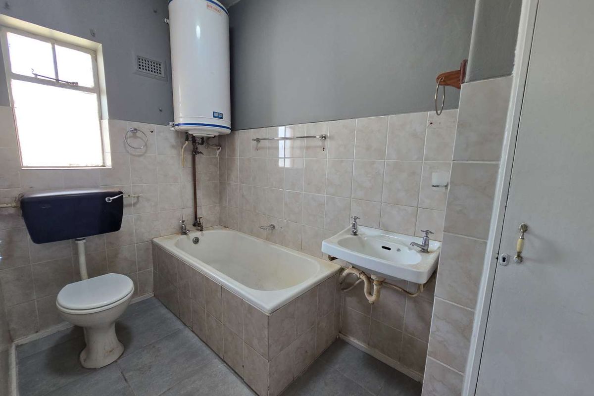 A dated bathroom featuring a bathtub, pedestal basin, and high-level cistern toilet, with exposed water heater plumbing and neutral wall tiling.