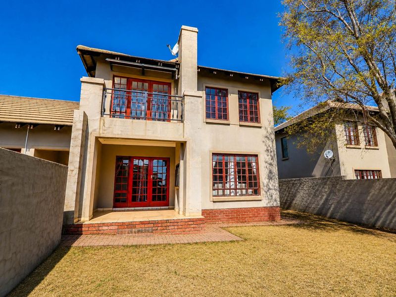 Rear view of a two-story house featuring a brick chimney, balcony with metal railing, tiled patio, and red-framed lattice windows overlooking a private garden.