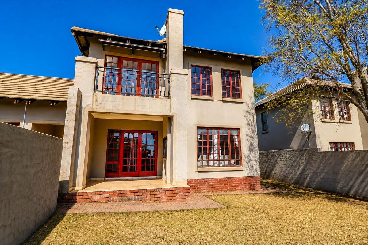 Rear view of a two-story house featuring a brick chimney, balcony with metal railing, tiled patio, and red-framed lattice windows overlooking a private garden.