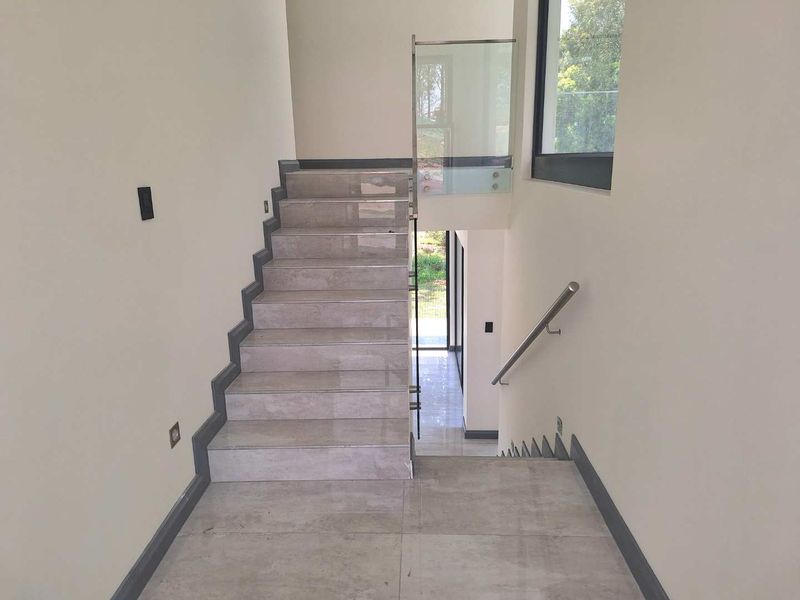A modern, well-lit hallway featuring a tiled staircase with a sleek glass balustrade and stainless steel handrail, finished with large format floor tiles.