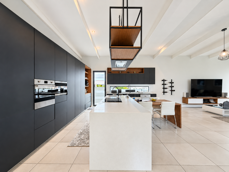 Modern open-plan kitchen and living area featuring sleek dark cabinetry, a large stone-topped island, integrated appliances, and high-quality tiled flooring throughout.