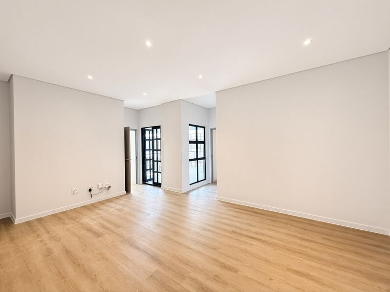 A bright, modern, and empty living area featuring neutral walls, light wood-look laminate flooring, and stylish black-framed glass doors leading to an outdoor space.