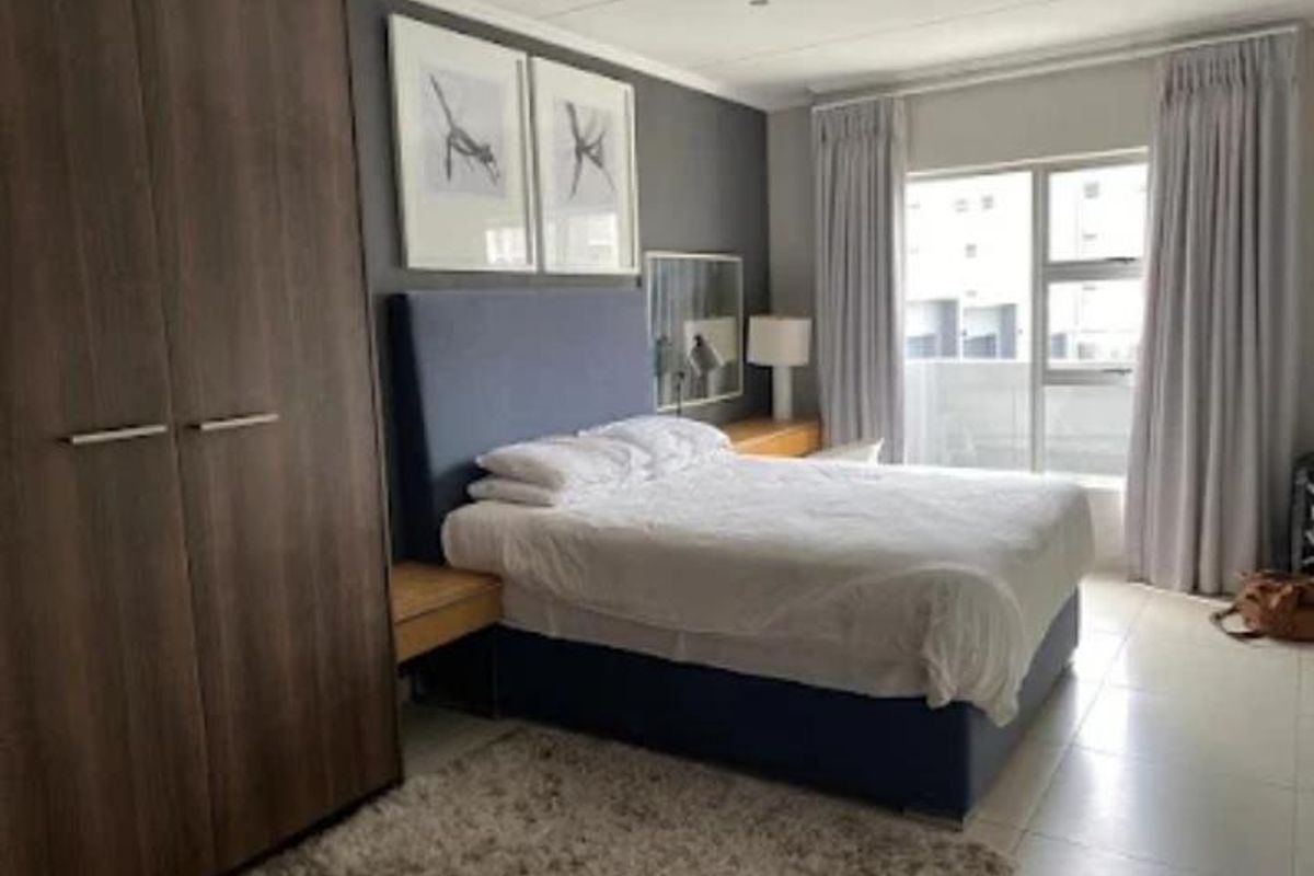 A well-lit bedroom featuring built-in wooden cabinetry, tiled flooring, and large windows with curtains providing natural light and access to a balcony.