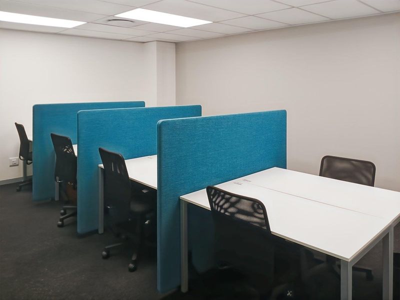A functional office space featuring rows of desks with integrated blue privacy partitions, suspended ceiling tiles, and carpeted flooring.