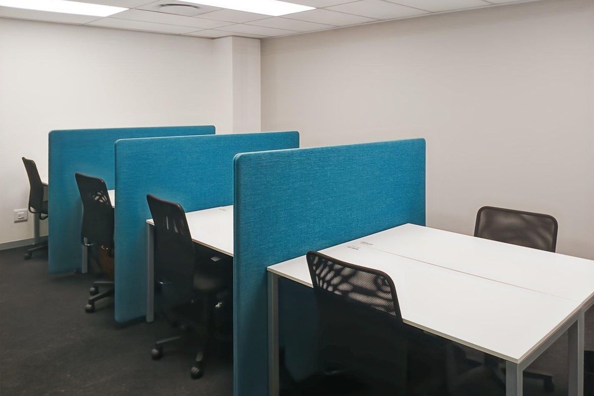 A functional office space featuring rows of desks with integrated blue privacy partitions, suspended ceiling tiles, and carpeted flooring.