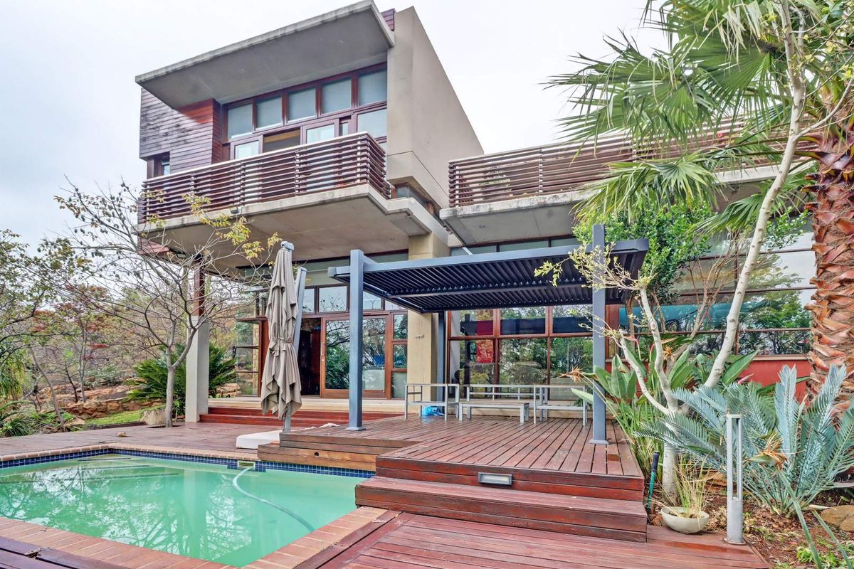 Rear exterior view featuring a modern multi-story residence with timber decking, a louvered pergola, and a swimming pool set in a landscaped garden.
