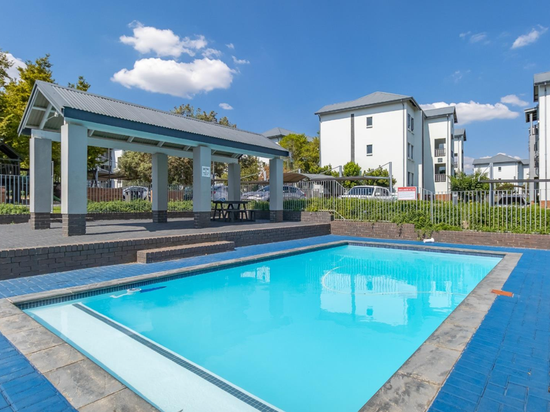 A well-maintained communal swimming pool area featuring a covered patio structure, brick paving, and secure perimeter fencing within an apartment complex.