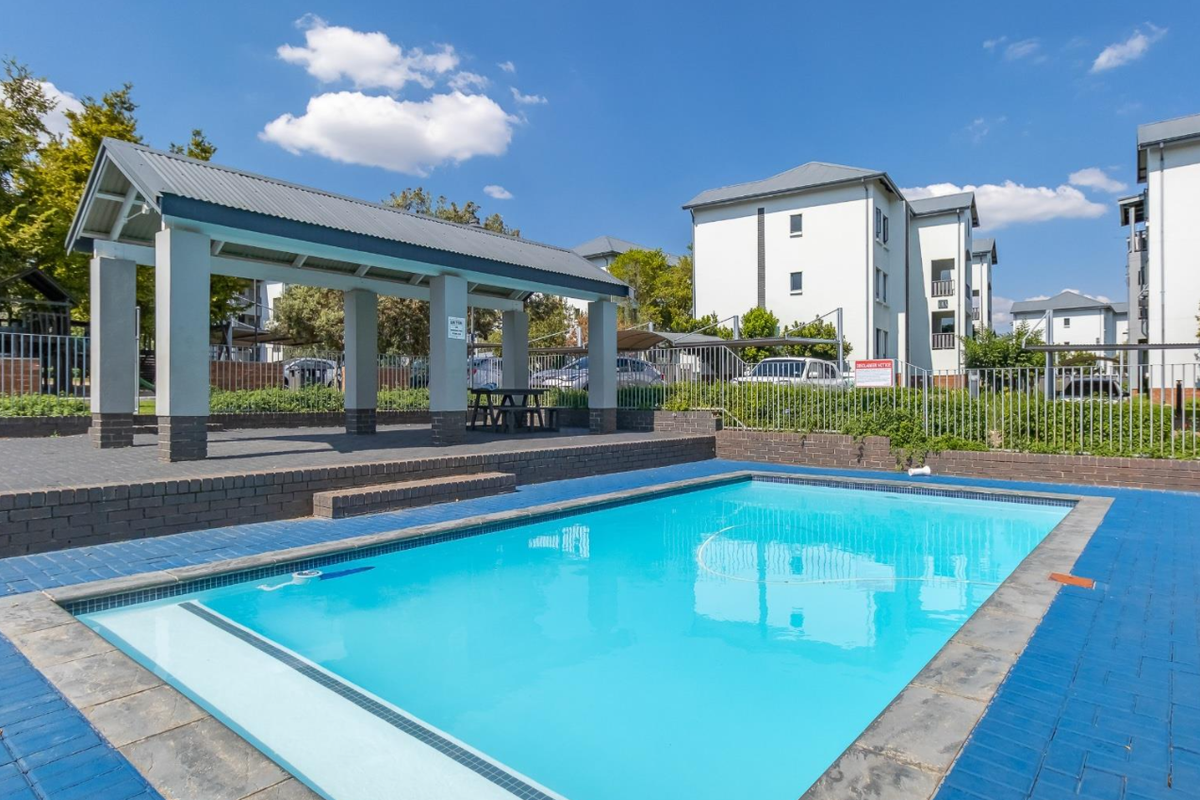A well-maintained communal swimming pool area featuring a covered patio structure, brick paving, and secure perimeter fencing within an apartment complex.