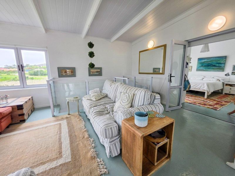 A bright living area featuring exposed beam ceilings, a glass balustrade, and direct access to an adjacent bedroom through a glass-paneled door.