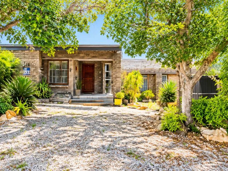 A charming single-story brick house featuring a gravel driveway, mature trees, and a welcoming timber front entrance.