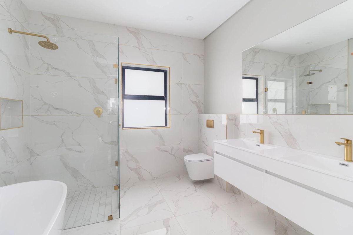 Modern bathroom featuring marble-look wall and floor tiles, a frameless glass shower, wall-mounted toilet, and a sleek double vanity with gold-toned fixtures.