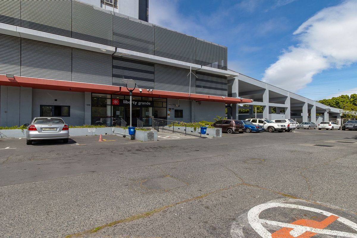 Exterior view of the Liberty Grande apartment building entrance, featuring a paved parking area, ramp access, and modern metal louvre architectural detailing.