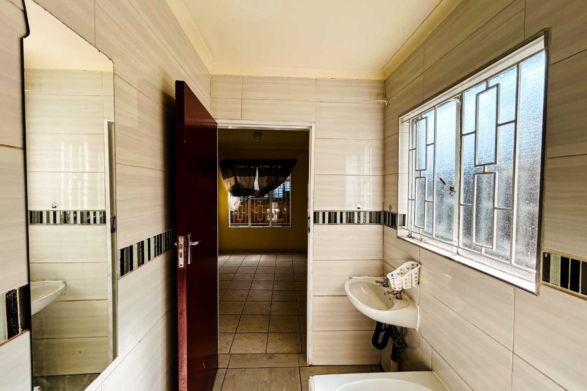A functional bathroom featuring tiled walls and flooring, a wall-mounted basin, and a window with security bars, leading into an adjacent room.